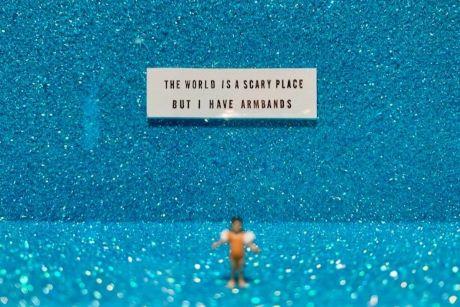 Human figurine with inflatable arm swim bands. Sign says "The World is a scary place but I have armbands"