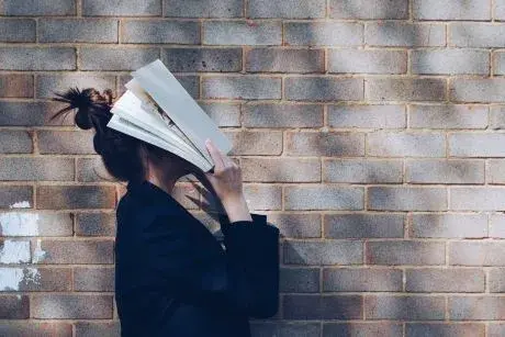 Person with bun on top of head wearing a black shirt standing in front of brick wall and holding an open book over their face.