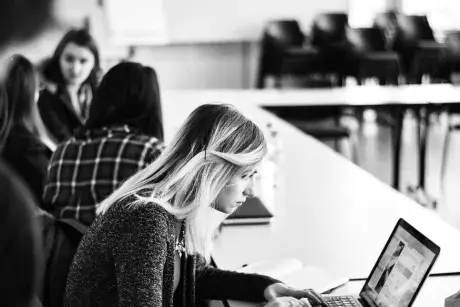 Female student on computer in classroom. Small group of students behind her talking.