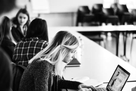 Female student on computer in classroom. Small group of students behind her talking.