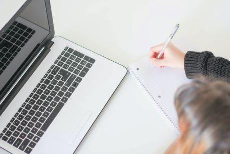 Woman writing in a notebook while seated in front of a laptop